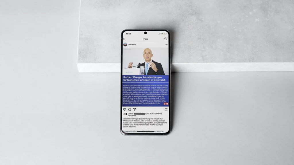 Smartphone liegt auf einer hellgrauen Oberfläche und zeigt einen Social-Media-Post mit Text und einem Redner vor Mikrofon