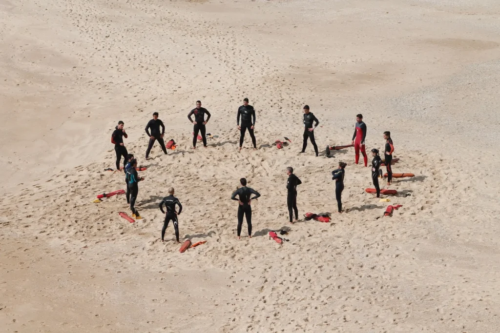Gruppe von Menschen in Neoprenanzügen steht im Kreis auf einem Sandstrand, umgeben von Rettungsbrettern.