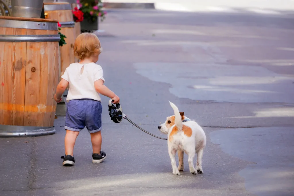 Kleines Kind geht auf einer Straße und führt einen Hund an der Leine, umgeben von Holzfässern und Blumen.