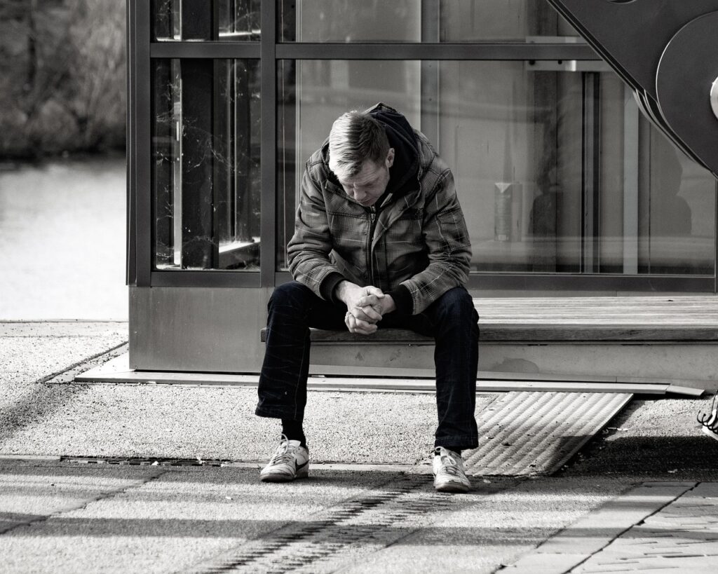 Schwarz weißes Bidl. Ein Mann sitzt vor einem Glasgebäude auf einer Betonbank und sieht zu Boden. Er ist Mitte 40, kurze Haare, dunkle Hose und hellere Jacke, mit weißen Turnschuhen. Er ist traurig / verzweifelt.