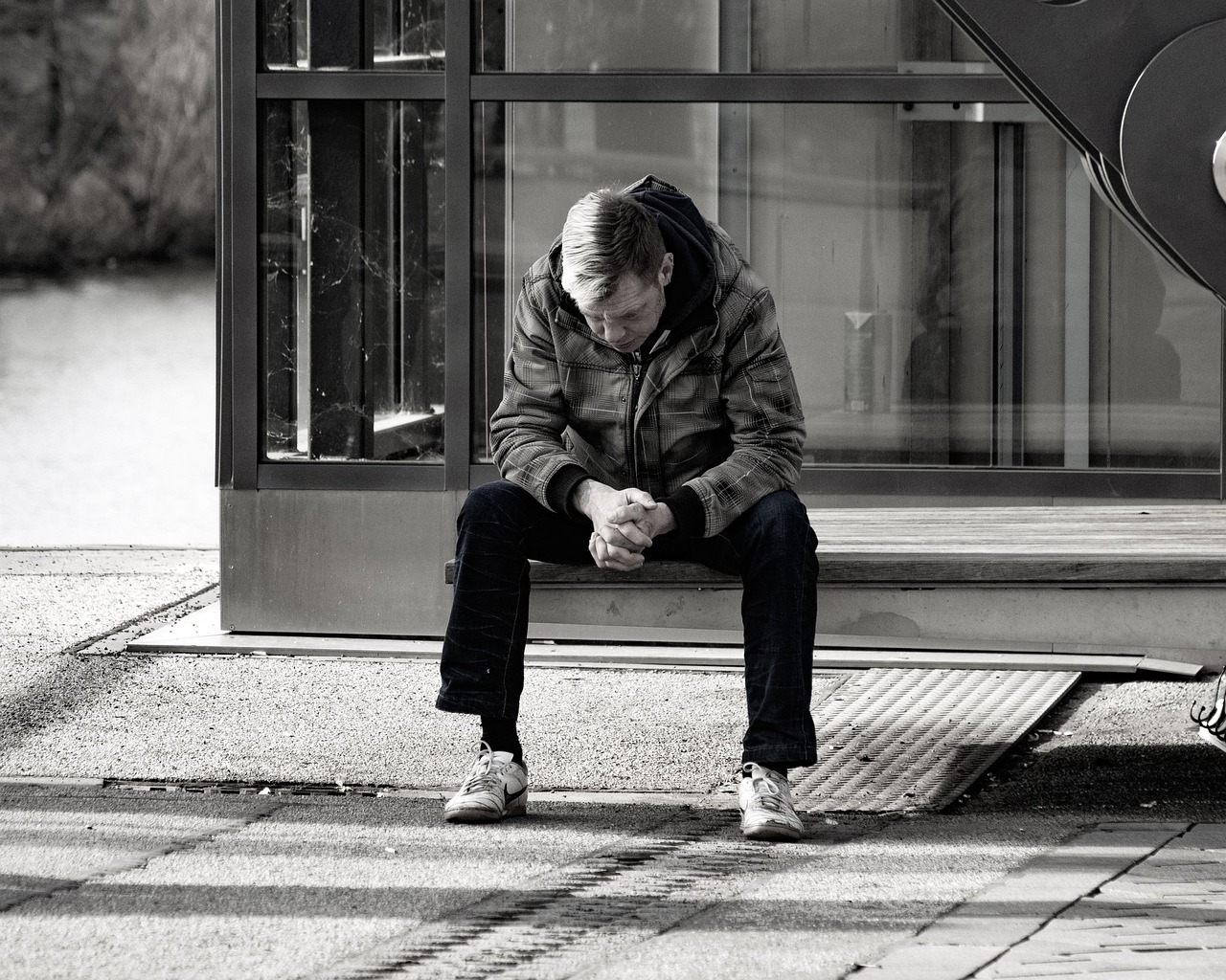 Schwarz weißes Bidl. Ein Mann sitzt vor einem Glasgebäude auf einer Betonbank und sieht zu Boden. Er ist Mitte 40, kurze Haare, dunkle Hose und hellere Jacke, mit weißen Turnschuhen. Er ist traurig / verzweifelt.
