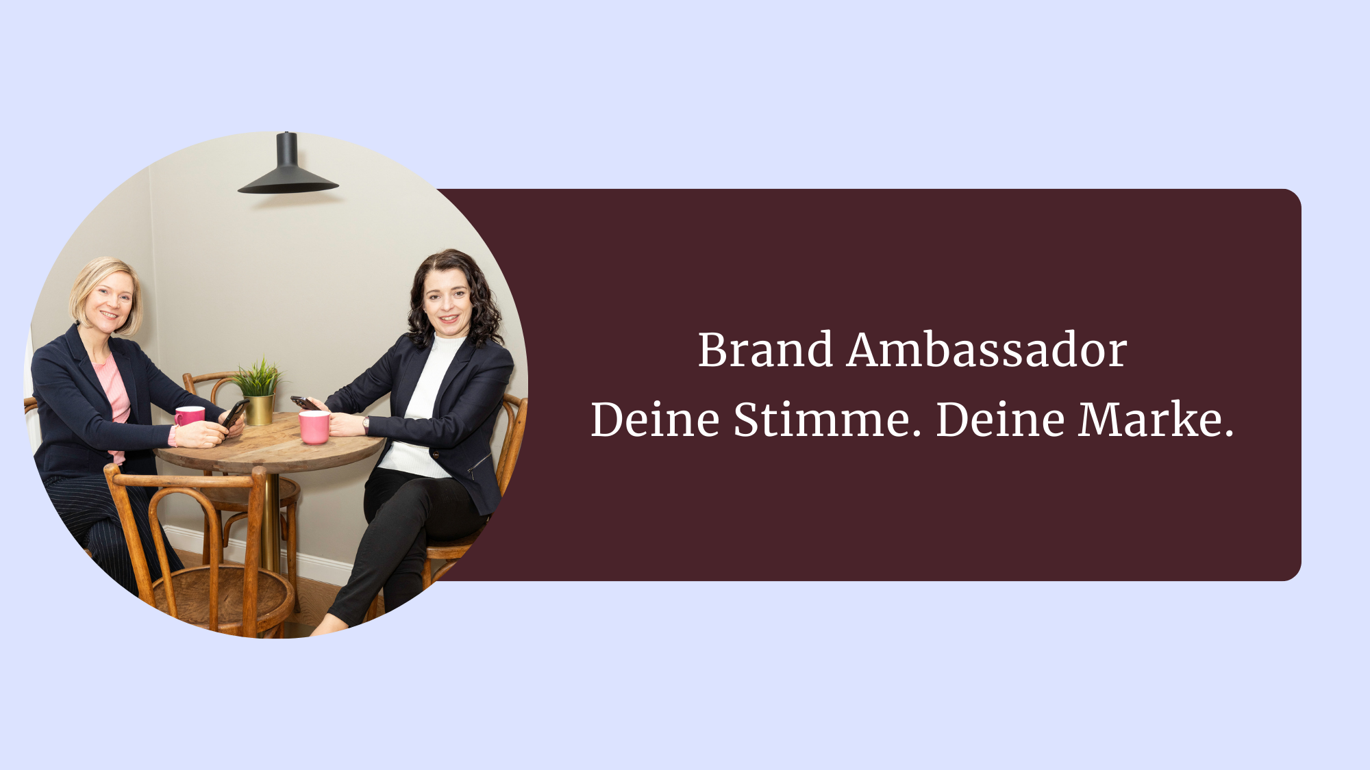Zwei schlanke Frauen in einem dunkelblauen Anzug, eine mit Rosa Shirt, die andere mit Weißem - sitzen gegenüber auf einem Tisch und schauen in die Kamera. Eine ist blond, kurzhaarig und die andere lange, dunkle gelockte Haare. Sie haben ein Handy in der Hand lächeln in die Kamera.