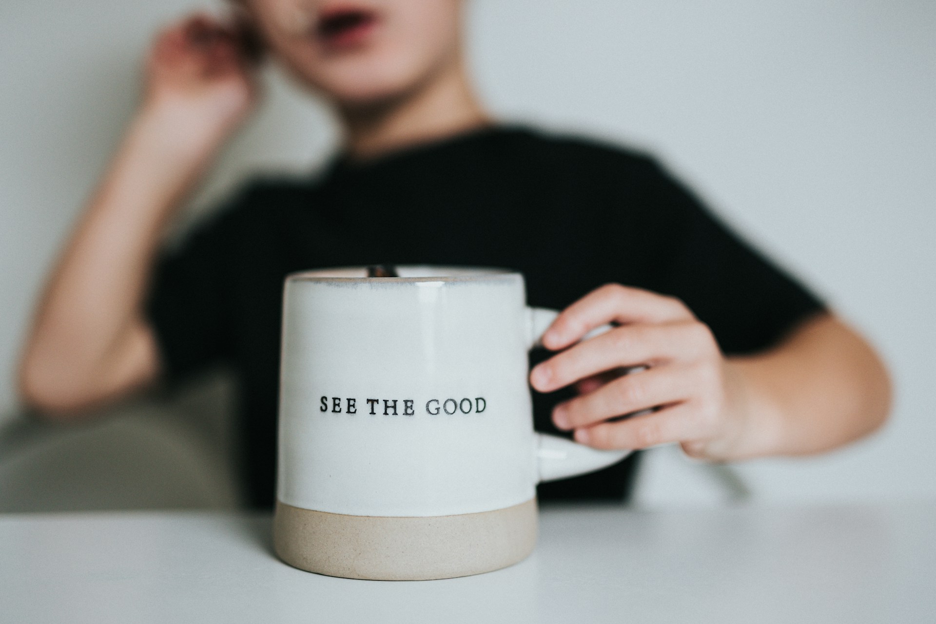 Eine Junge in schwarzen T-Shirt, kurzarmig, im Hintergrund hält eine Tasse in der Hand auf der steht - see the good.