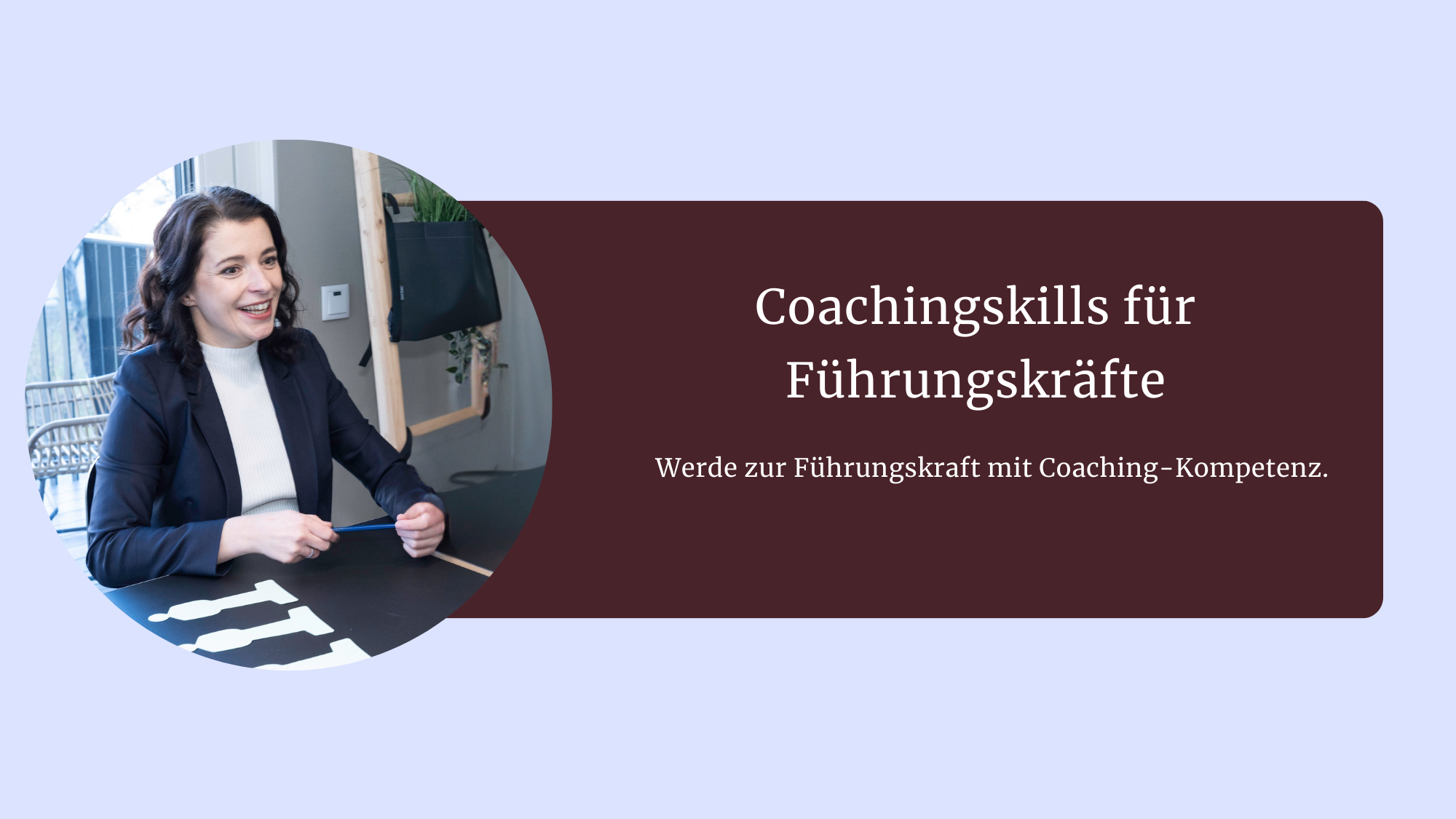 Junge Frau mit dunklen, gelockten Haaren, lächelt und hält einen Bleistift in der Hand. Sie hat ein dunkles Sakko an und einen weißen Rollkragenpullover. Sie sitzt an einem dunklen Tisch, Fenster im Hintergrund.