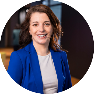 Im Gespräch mit Martina Ernst Junge Frau mit dunklen Haaren, lächelt in die Kamera. Sie ist in einem Büro und trägt ein blitzblaues Sakko und ein weißes Top darunter.