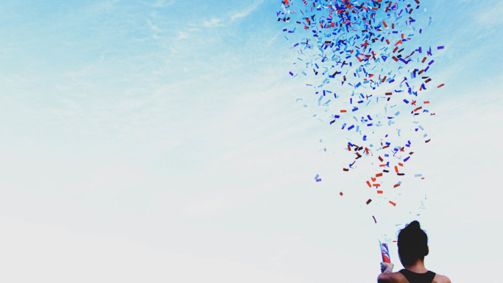 Eine junge Frau mit dunklen Haar schaut in den blauen Himmel mit einer Konfettikanone in der Hand.
